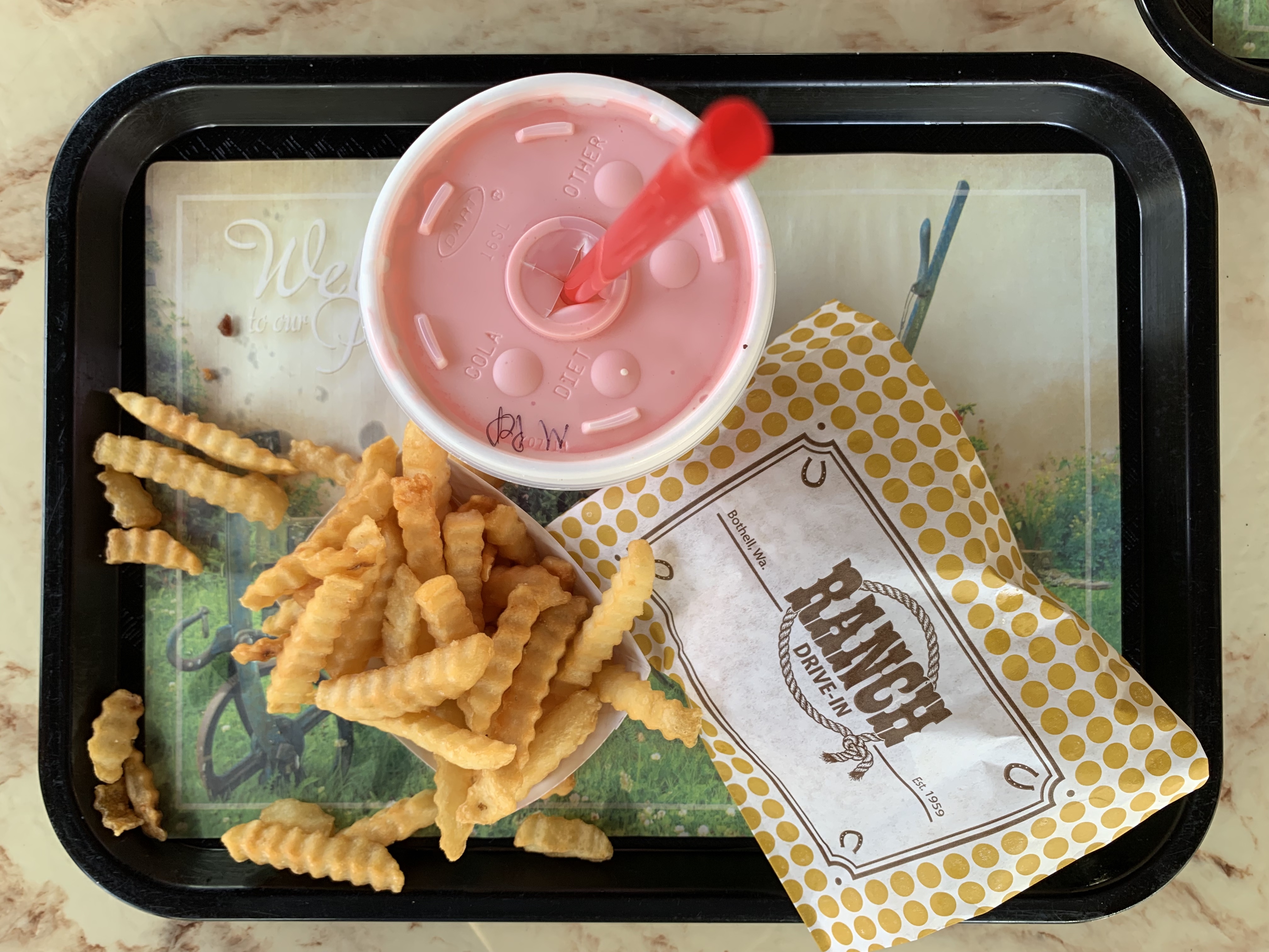 Tray of food from ranch drive-in. There's a pink milkshake, fries, and a burger wrapped on the green tray.