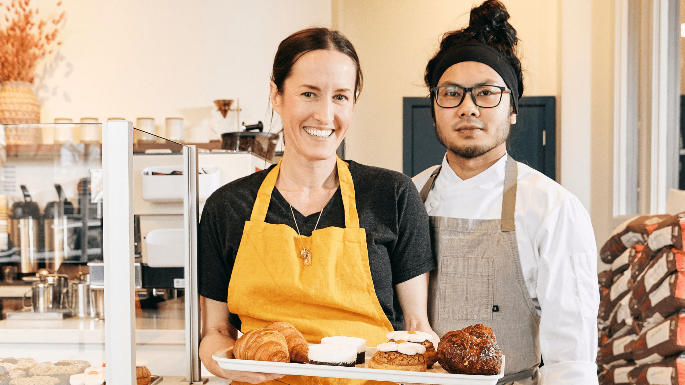 Two people in a bakery. One of them is wearing a yellow apron and holding up a tray of pastries.